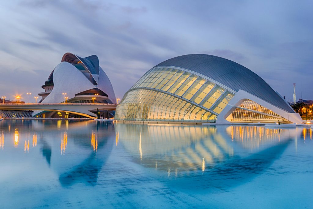 Futuristic curved glass and white concrete buildings illuminated at dusk, reflected in calm blue water