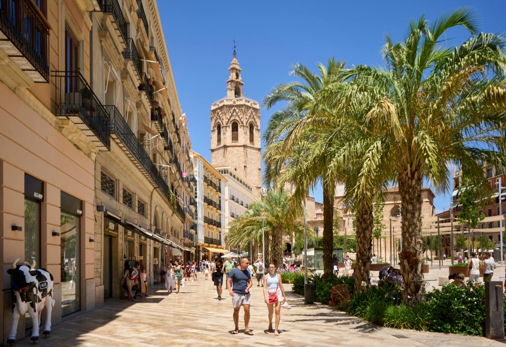 Sunny pedestrian street with palm trees, historic stone tower, and people walking between classic buildings