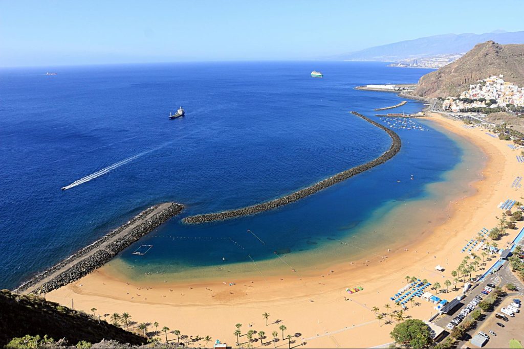 A wide panoramic view of a golden sandy beach with calm blue ocean water, curved breakwaters, small boats, and a coastal town set against mountains under a clear sky.