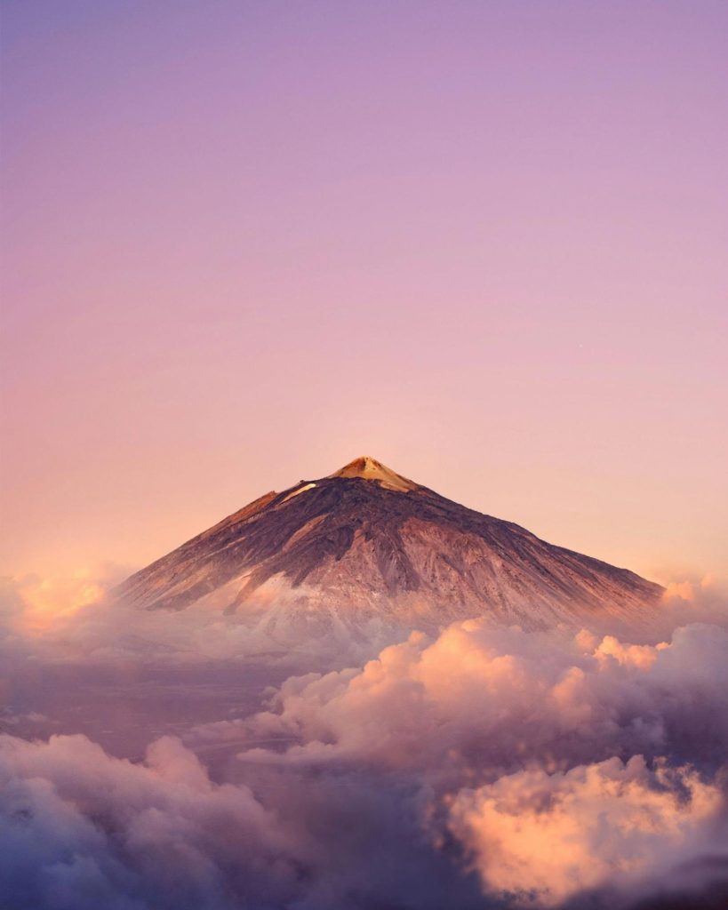 Snow-capped mountain peak rising above soft pink and golden clouds at sunset.