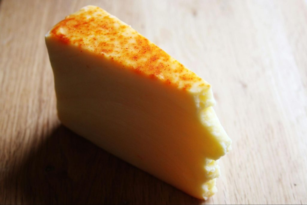 A wedge of semi-hard cheese sprinkled with red spices on top, placed on a wooden surface.